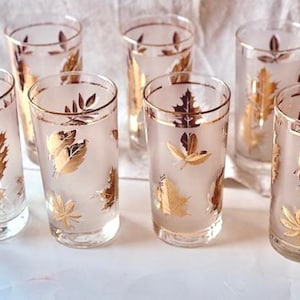 May include: Set of seven vintage highball glasses. Each glass features a frosted finish with gold leaf designs. The rims and leaf details are accented with gold. The glasses are cylindrical and sit on a white surface.