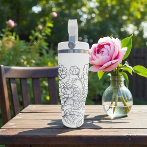 May include: White insulated tumbler with a black floral pattern. The tumbler has a handle and a silver rim. A pink rose in a glass vase sits on a wooden table next to the tumbler.