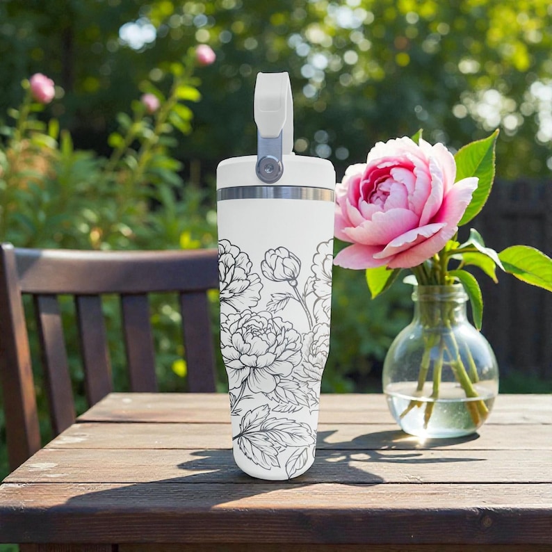 May include: White insulated tumbler with a black floral pattern. The tumbler has a handle and a silver rim. A pink rose in a glass vase sits on a wooden table next to the tumbler.
