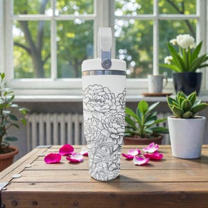 May include: A white insulated travel mug featuring a black and white floral pattern. It has a handle and a silver rim. Pink rose petals are scattered on a wooden table. The background includes plants and a window.