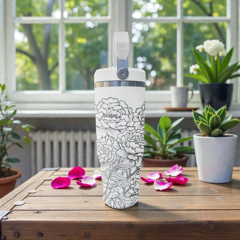 May include: A white insulated travel mug featuring a black and white floral pattern. It has a handle and a silver rim. Pink rose petals are scattered on a wooden table. The background includes plants and a window.