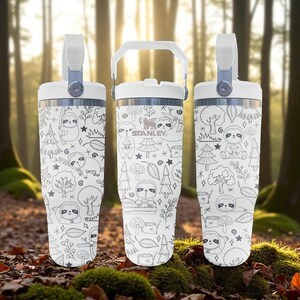 May include: Three white Stanley tumblers with a handle and a lid. Each tumbler is decorated with a black and white doodle design featuring sloths, trees, and mushrooms. The Stanley logo is visible on one of the tumblers.