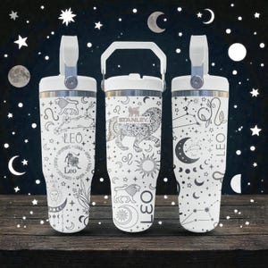 May include: Three white Stanley tumblers with handles and lids, each featuring black line art designs related to the Leo zodiac sign. The patterns include lions, suns, moons, stars, constellations, and the text Leo, arranged in various celestial motifs.