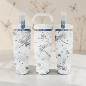 May include: Three white Stanley tumblers with a dragonfly and constellation design. Each tumbler has a white handle and lid, with a silver accent. The dragonfly design is in black and white. The Stanley logo is visible on the center tumbler.