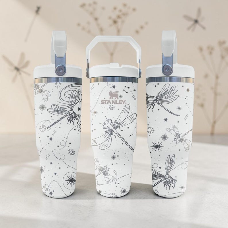 May include: Three white Stanley tumblers with a dragonfly and constellation design. Each tumbler has a white handle and lid, with a silver accent. The dragonfly design is in black and white. The Stanley logo is visible on the center tumbler.