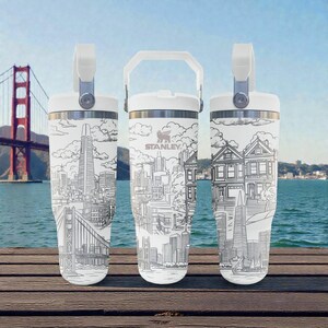 Puede incluir: Tres vasos aislados blancos con asa, con un dibujo lineal en blanco y negro del horizonte de San Francisco, incluyendo el Golden Gate Bridge y la Transamerica Pyramid. El logotipo de Stanley es visible.