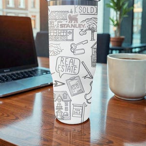 K&ouml;nnte beinhalten: Wei&szlig;er Edelstahlbecher mit schwarzem Strichdesign mit H&auml;usern, Schl&uuml;sseln und den W&ouml;rtern "REAL ESTATE" und "SOLD". Der Becher tr&auml;gt auch den Markennamen "STANLEY". Er steht auf einem Holztisch.