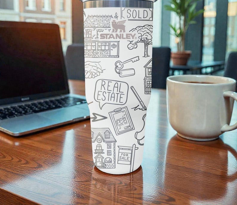 K&ouml;nnte beinhalten: Wei&szlig;er Edelstahlbecher mit schwarzem Strichdesign mit H&auml;usern, Schl&uuml;sseln und den W&ouml;rtern "REAL ESTATE" und "SOLD". Der Becher tr&auml;gt auch den Markennamen "STANLEY". Er steht auf einem Holztisch.