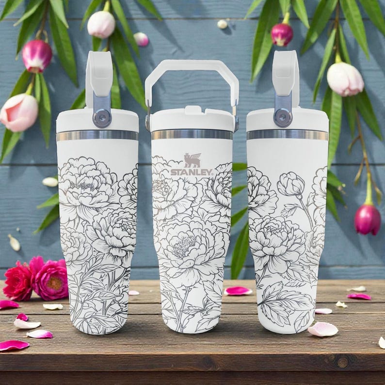 May include: Three white Stanley travel mugs with floral patterns. Each mug has a handle and a lid. The mugs are decorated with black and white peony flowers. The Stanley logo is visible on the center mug.