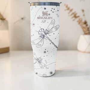 May include: White Stanley tumbler with a dragonfly and constellation design. The tumbler features a silver rim and the Stanley logo. The dragonfly and constellation design is in gray. The tumbler is cylindrical and appears to be made of stainless steel.
