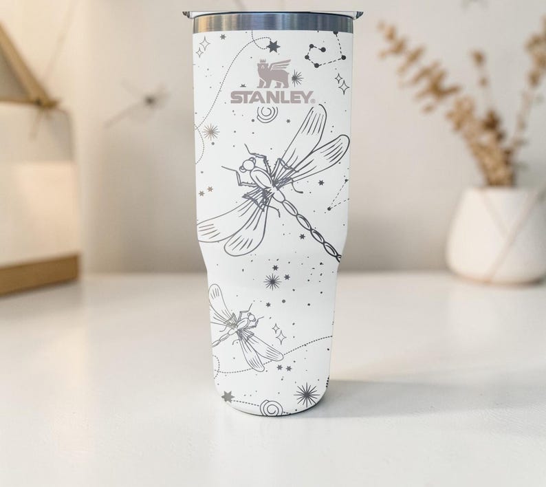 May include: White Stanley tumbler with a dragonfly and constellation design. The tumbler features a silver rim and the Stanley logo. The dragonfly and constellation design is in gray. The tumbler is cylindrical and appears to be made of stainless steel.