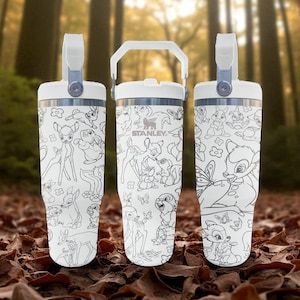 May include: White insulated tumbler with a black and white Bambi and forest creatures illustration. The tumbler has a white handle and lid, with the brand name "Stanley" printed on the side. The background shows a forest setting.