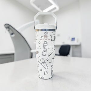 May include: White Stanley tumbler with a handle, featuring a dental-themed pattern in black. The design includes toothbrushes, toothpaste, and other dental-related icons. The Stanley logo is visible. The tumbler is on a white surface.