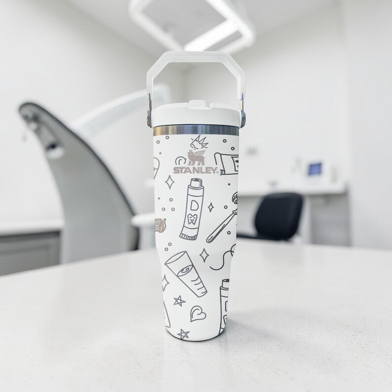 May include: White Stanley tumbler with a handle, featuring a dental-themed pattern in black. The design includes toothbrushes, toothpaste, and other dental-related icons. The Stanley logo is visible. The tumbler is on a white surface.