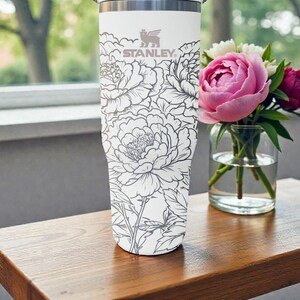 May include: A white Stanley travel mug with a grey floral pattern. The mug has the Stanley logo and is placed beside a vase of pink and white flowers on a wooden surface.