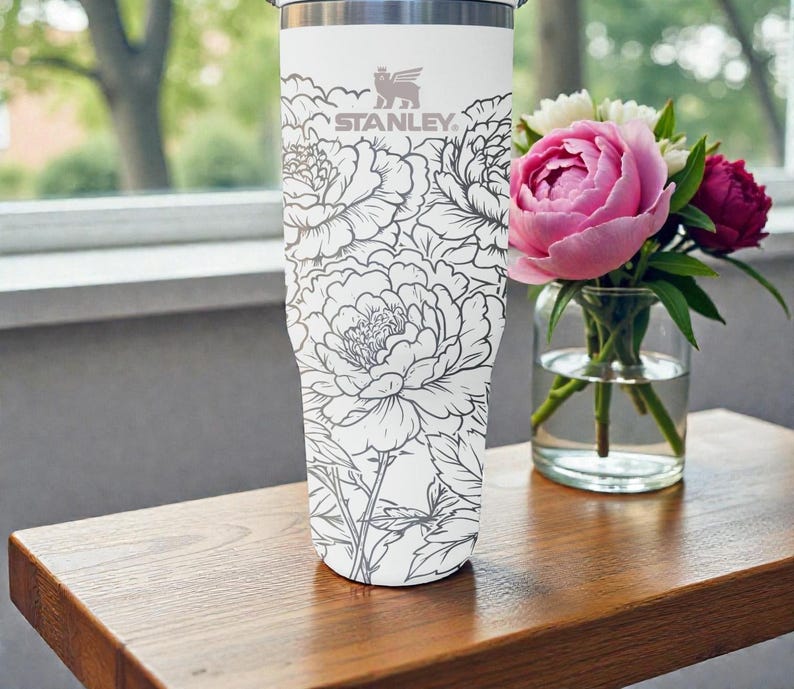 May include: A white Stanley travel mug with a grey floral pattern. The mug has the Stanley logo and is placed beside a vase of pink and white flowers on a wooden surface.