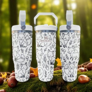 May include: Three white Stanley travel mugs with a black and white fox and woodland pattern. Each mug has a white handle and lid. The mugs are on a bed of moss with acorns and leaves.