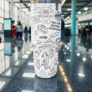 Puede incluir: Un vaso de viaje Stanley blanco con un dise&ntilde;o en blanco y negro con tem&aacute;tica de viajes. El dise&ntilde;o incluye aviones, equipaje y el texto "Time for Another Adventure!" y "Stanley". El vaso es cil&iacute;ndrico y parece ser de acero inoxidable.