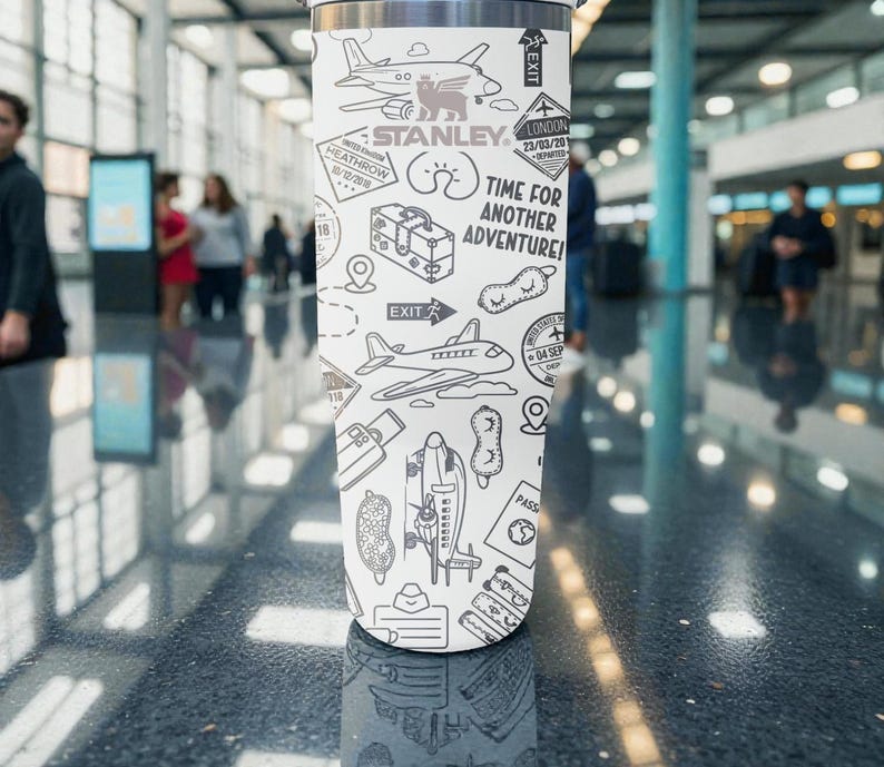 Puede incluir: Un vaso de viaje Stanley blanco con un dise&ntilde;o en blanco y negro con tem&aacute;tica de viajes. El dise&ntilde;o incluye aviones, equipaje y el texto "Time for Another Adventure!" y "Stanley". El vaso es cil&iacute;ndrico y parece ser de acero inoxidable.