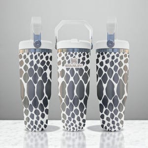 May include: Three stainless steel tumblers with a white and dark grey animal print pattern. Each tumbler features a white lid with a sturdy handle and a metal band. The centre tumbler displays the Stanley logo in white on the front surface.