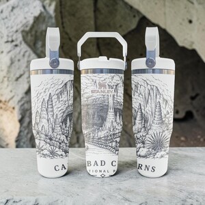 May include: Three white Stanley tumblers featuring a black and white line art illustration of Carlsbad Caverns National Park. The design depicts cave formations, stalactites, and a walking path. The text reads Carlsbad Caverns National Park.