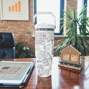 K&ouml;nnte beinhalten: Ein wei&szlig;-silberner isolierter Becher mit wei&szlig;em Griff, verziert mit einem schwarz-wei&szlig;en Doodle-Design zum Thema Immobilien. Das Design umfasst H&auml;user, Schl&uuml;ssel und die W&ouml;rter "SOLD" und "REAL ESTATE".
