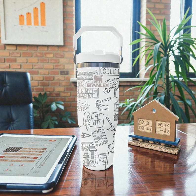 K&ouml;nnte beinhalten: Ein wei&szlig;-silberner isolierter Becher mit wei&szlig;em Griff, verziert mit einem schwarz-wei&szlig;en Doodle-Design zum Thema Immobilien. Das Design umfasst H&auml;user, Schl&uuml;ssel und die W&ouml;rter "SOLD" und "REAL ESTATE".