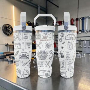 Op de afbeelding: Drie witte Stanley-bekers met een witte handgreep en een zwart lijnpatroon met auto-onderdelen zoals motoren, zuigers, moersleutels en auto's. De tekst bevat "Built Not Bought", "Mechanic Mode On", "Born to Drive" en "Eat Sleep Race Repeat".