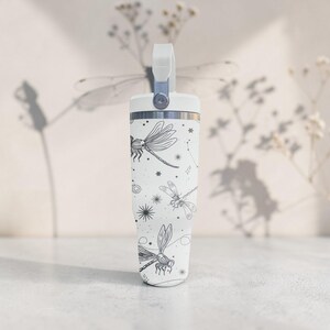May include: White tumbler with a handle, featuring a black dragonfly and constellation pattern. The tumbler has a silver rim and a white handle. The design includes stars and celestial elements. The tumbler is designed for cold beverages.