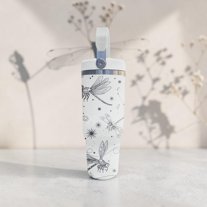 May include: White tumbler with a handle, featuring a black dragonfly and constellation pattern. The tumbler has a silver rim and a white handle. The design includes stars and celestial elements. The tumbler is designed for cold beverages.