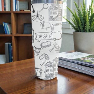 K&ouml;nnte beinhalten: Ein wei&szlig;er Edelstahlbecher mit einem schwarz-wei&szlig;en Immobilien-Design. Das Design umfasst Illustrationen von H&auml;usern, Schl&uuml;sseln, Geld und den Text "FOR SALE". Der Becher steht auf einem Holztisch.
