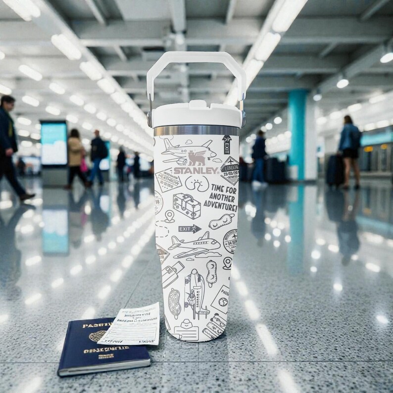 Puede incluir: Un termo de viaje blanco y plateado con asa, con ilustraciones en blanco y negro con tem&aacute;tica de viajes y el texto "TIME FOR ANOTHER ADVENTURE!". Un pasaporte y documentos de viaje est&aacute;n en primer plano. El termo est&aacute; sobre un suelo reflectante.