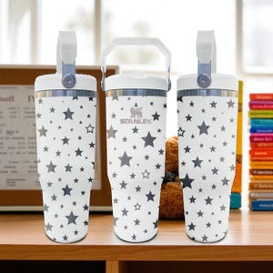 May include: Three white stainless steel tumblers with a repeating pattern of dark gray stars. Each tumbler features a white lid with a handle, and the center tumbler displays the Stanley brand logo in dark gray text on the upper portion.