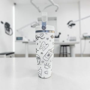 May include: A white insulated tumbler with a handle, featuring a pattern of dental-themed doodles in black. The design includes teeth, hearts, stars, and toothpaste tubes. The tumbler is standing on a white surface.