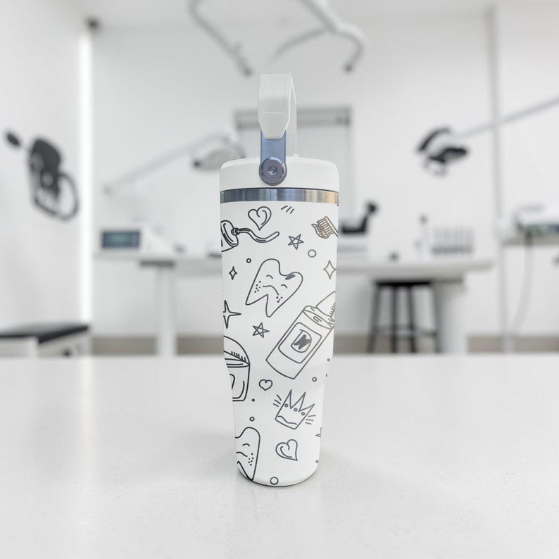 May include: A white insulated tumbler with a handle, featuring a pattern of dental-themed doodles in black. The design includes teeth, hearts, stars, and toothpaste tubes. The tumbler is standing on a white surface.