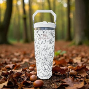 May include: White insulated tumbler with a handle, featuring a koala and leaf pattern. The tumbler has a white lid and handle, with the brand name "STANLEY" printed on the front. The background is a blurred forest scene with fallen leaves.