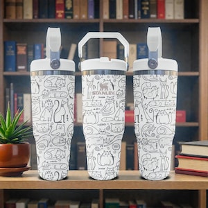May include: Three white stainless steel tumblers with a repeating black line art pattern of various cats and fish bones. Each tumbler features a white lid with a handle and the Stanley logo printed on the centre unit in black ink.