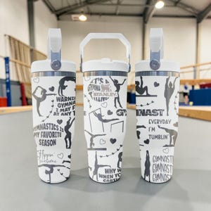 Pode incluir: Três copos brancos de aço inoxidável com tampas e alças brancas, apresentando gráficos pretos com tema de ginástica e textos incluindo "Gymnastics is my favorite season", "Flippin' Awesome", "Everyday I'm tumblin'" e "It's a gymna thing".