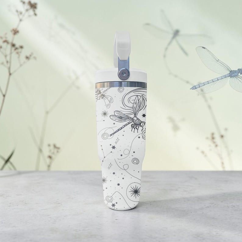 May include: White insulated tumbler with a handle and a black dragonfly design. The tumbler features a screw-on lid and a handle. The design includes dragonflies, stars, and celestial elements.