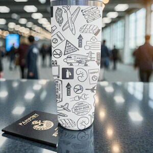 Puede incluir: Un vaso de viaje blanco con borde de acero inoxidable, cubierto con dibujos lineales negros de aviones, sellos de pasaporte e iconos de viaje. Un pasaporte negro con letras doradas est&aacute; delante. El vaso mide aproximadamente 23 cm de alto.
