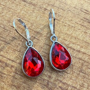 May include: A pair of silver-toned drop earrings featuring large, faceted, teardrop-shaped red gemstones. The earrings have a lever-back closure and are displayed on a wooden surface. The gemstones have a brilliant, sparkling appearance.