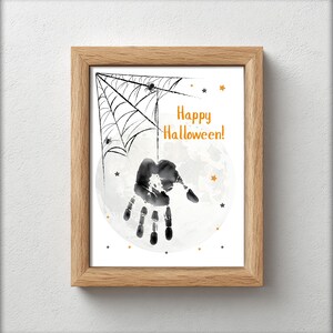 Pode incluir: Uma impressão de Halloween emoldurada com uma impressão de mão pendurada em uma teia de aranha contra um fundo de lua cheia. As palavras "Happy Halloween!" estão em laranja. A moldura é de madeira marrom clara.