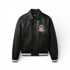 May include: Black leather jacket with a zip-up front, collar, and a patch on the chest. The patch features a design with red, white, and blue colors. The jacket has white stripes on the cuffs and waist. The brand name "AVIREX" is visible.