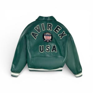 May include: A dark green leather jacket with the word "AVIREX" arched across the upper back. Below the word "AVIREX" is a patch with the American flag and the words "ALL AMERICANS ESTABLISHED 1975". The word "USA" is below the patch.