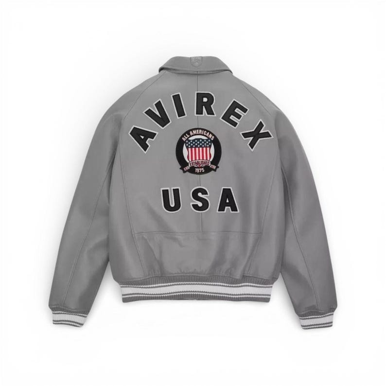 May include: A gray leather jacket with the word "AVIREX" arched across the back, above a circular emblem and the word "USA". The emblem features a red, white, and blue design with the words "ALL AMERICANS" and "ESTABLISHED 1975". The jacket has a ribbed hem and cuffs.