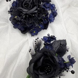 May include: Two floral arrangements featuring black fabric roses, dark blue accent flowers, and black beaded accents. One arrangement is a cluster, the other is a boutonniere with a green stem. The background is a white, textured fabric.