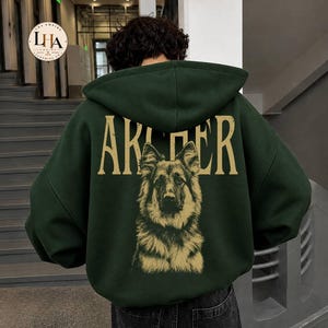 May include: A dark green hooded sweatshirt featuring a large, tan-colored graphic of a German Shepherd dog on the back. Above the dog, the text "ARCHER" is printed in a distressed, tan-colored font. The hoodie has a hood and long sleeves.