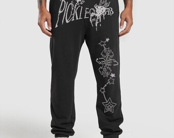 May include: Black sweatpants with a drawstring waist and white graphic text that reads "PICKLE LEGS". A constellation design with stars and other shapes is printed down the right leg. The sweatpants are paired with white sneakers.
