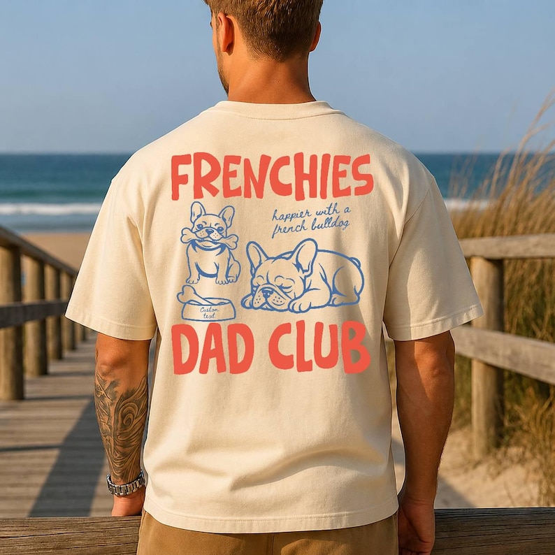 May include: Cream-colored t-shirt with the words "FRENCHIES" and "DAD CLUB" in red. The shirt features blue illustrations of French bulldogs and the phrase "happier with a french bulldog".