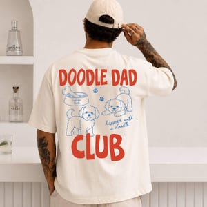 Op de afbeelding: Crèmekleurig T-shirt met de woorden "DOODLE DAD CLUB" in rood en blauw. Het shirt heeft blauwe pootafdrukken en doodle-hondenillustraties. De zin "happier with a doodle" staat er ook op.